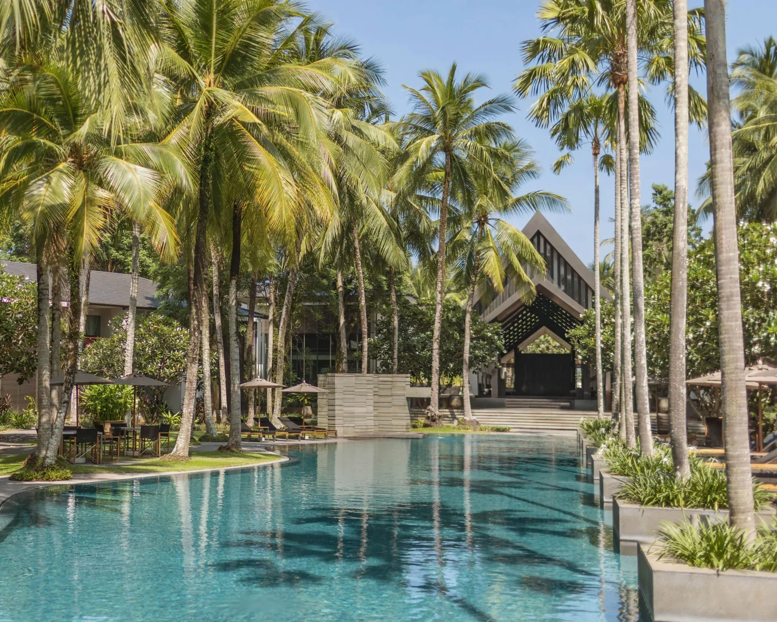 Лагунный бассейн отеля Twinpalms Surin Beach Phuket среди высоких пальм с шезлонгами и входным павильоном с остроконечной крышей