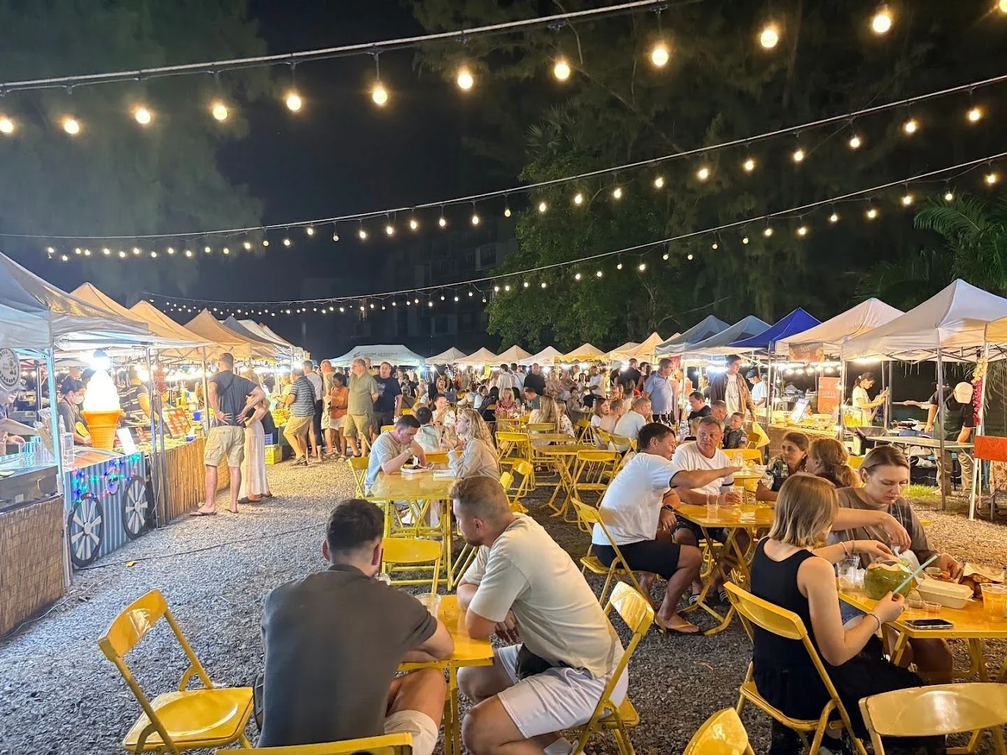 Заполненные столики между двумя рядами палаток Laguna Lifestyle Night Market под гирляндами вечерних ламп.
