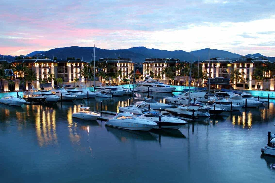Причалы Royal Phuket Marina на закате: яхты у понтонов, подсвеченные здания марина-виллидж и горы на горизонте.