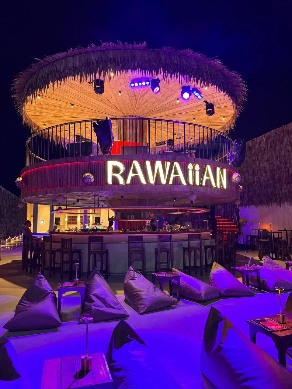 Круглый бар Rawaiian ночью: соломенная крыша, светящаяся вывеска, барные стулья и кресла-мешки в фиолетовой подсветке