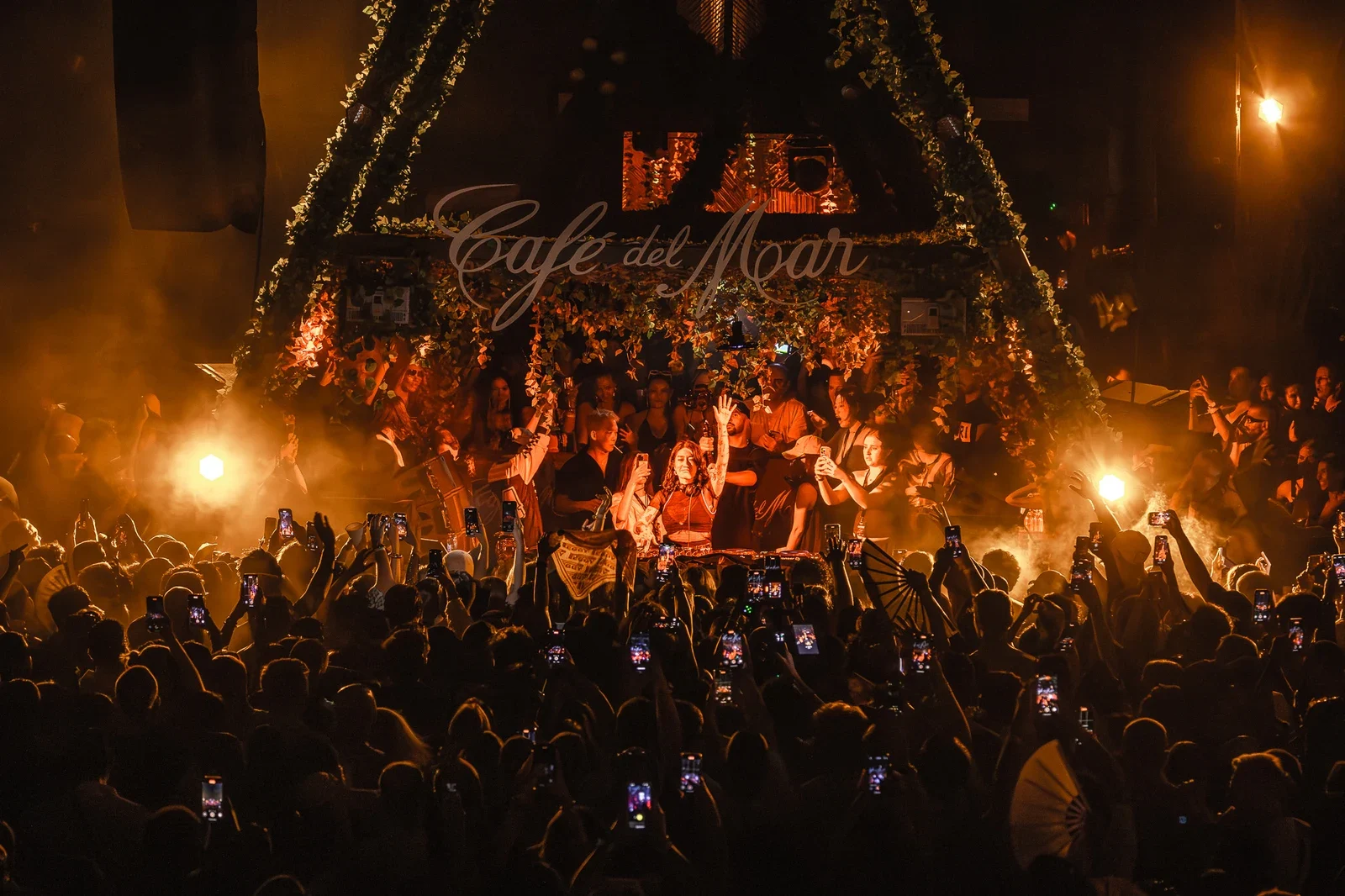 Ночной сет диджея под вывеской Café del Mar в плюще, над толпой с телефонами поднятые руки, дым и тёплый свет