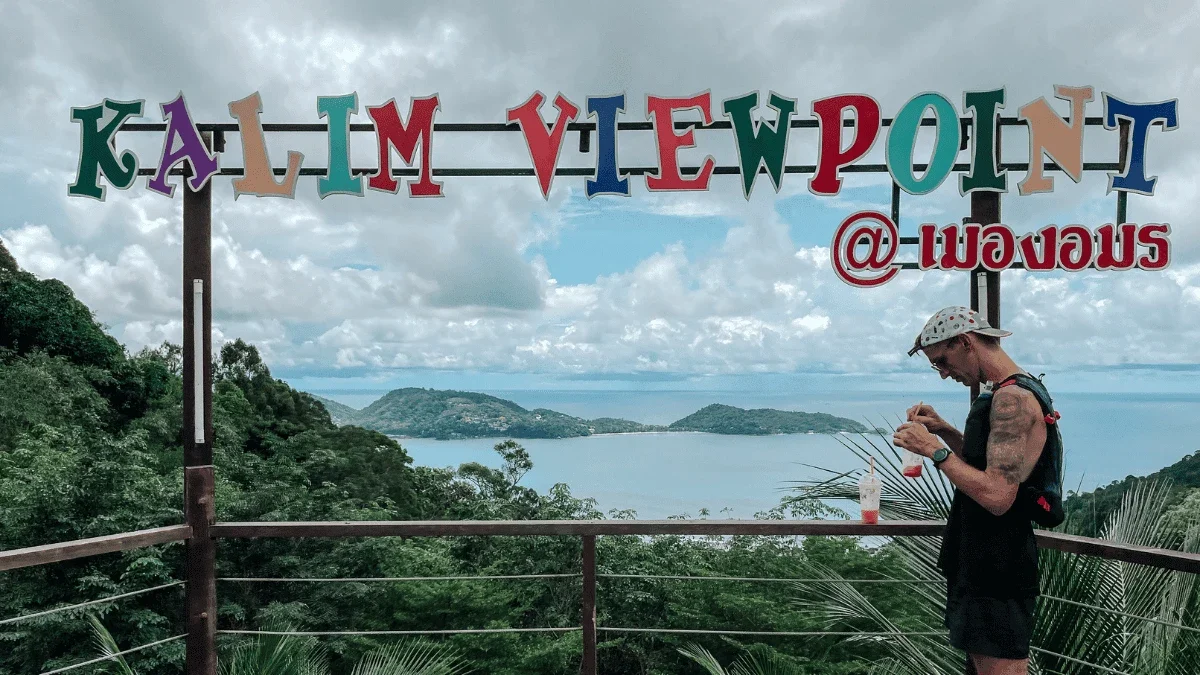 Человек на смотровой площадке Калим под разноцветной вывеской KALIM VIEWPOINT, за ним бухта и зелёные холмы Пхукета.