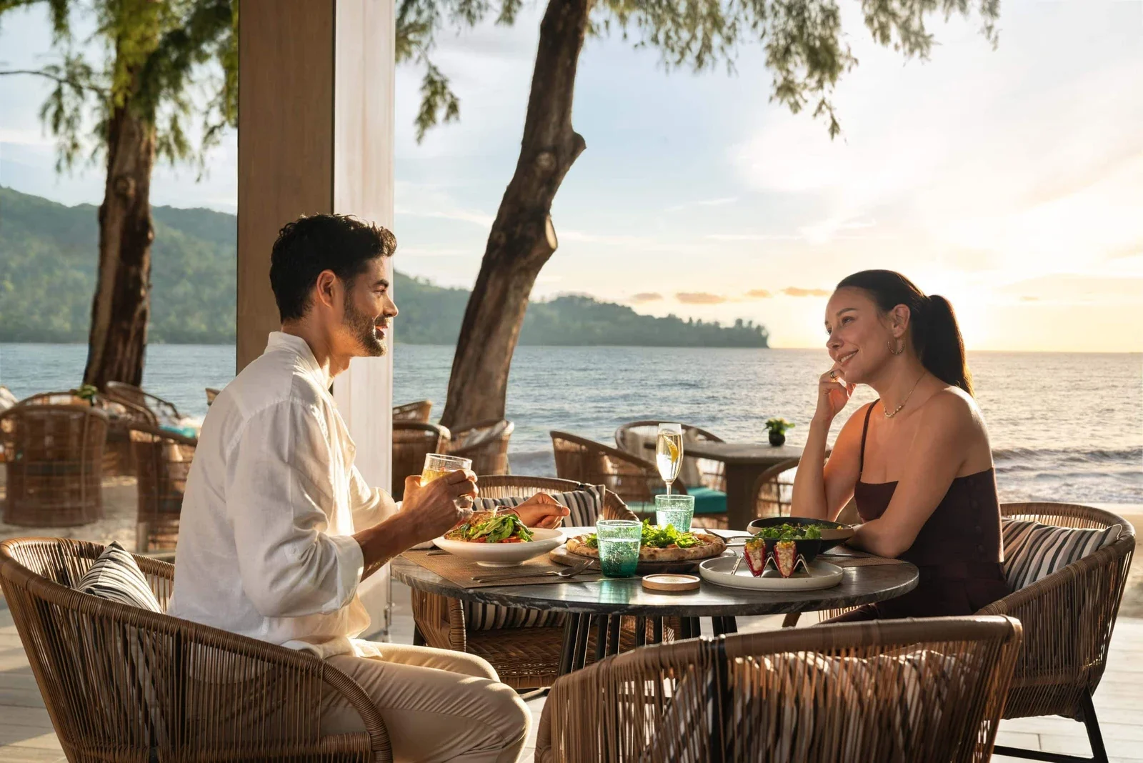 Пара обедает за столиком на террасе Pine Beach Bar, позади море, лесистый берег и закатное небо