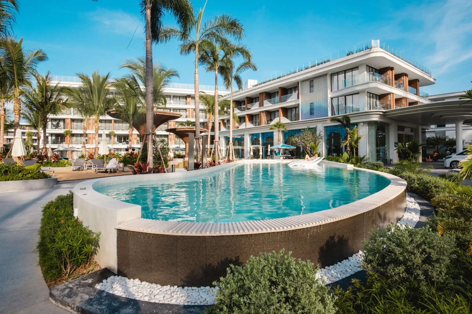 Главный корпус Kora Beach Resort с въездом под навесом и изогнутым бассейном в саду