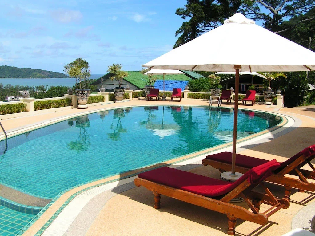 Бассейн Prince Edouard Apartments & Resort Phuket с красными шезлонгами, белым зонтом и видом на залив за ограждением.