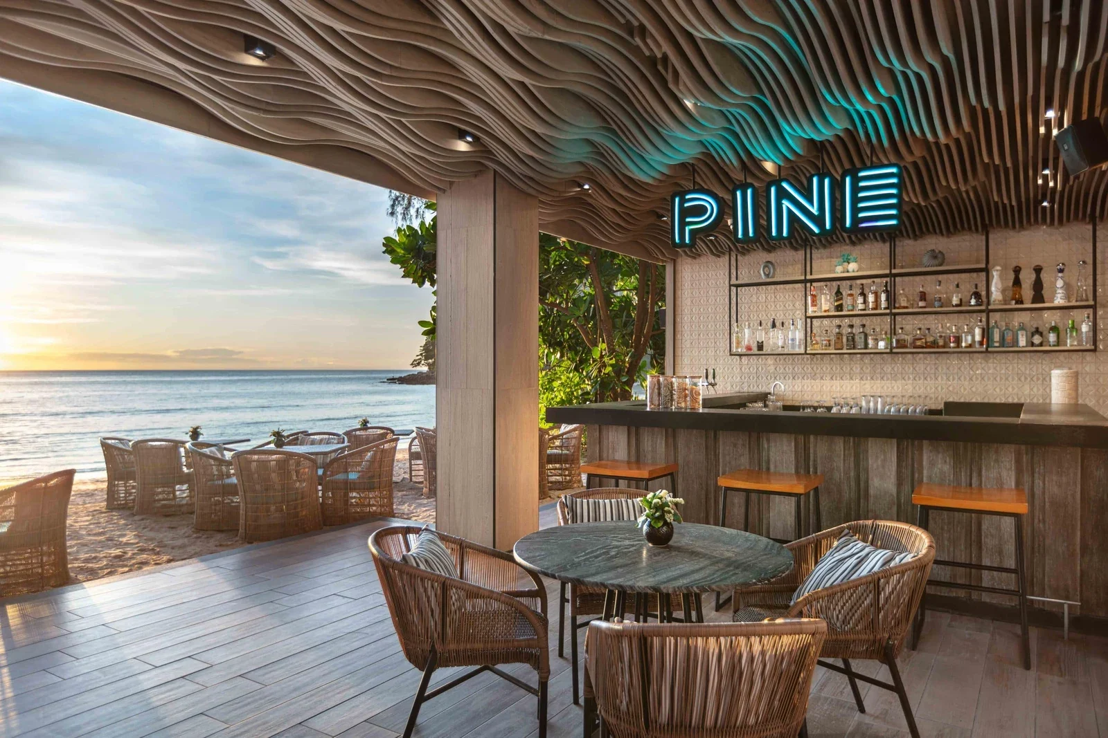 Барная стойка Pine Beach Bar под волнистым деревянным потолком с неоновой вывеской PINE, столики на террасе у моря