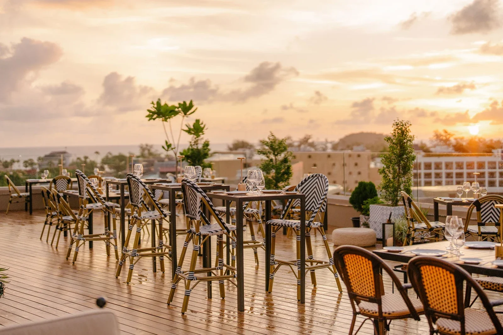 Открытая терраса Moon Terrace Lounge на закате: столы с плетёными стульями на деревянном настиле и вид на побережье Каты