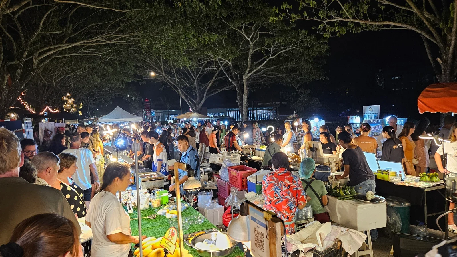 Прилавки и посетители ночного рынка Fun Friday Avenue под деревьями в Бангтао.