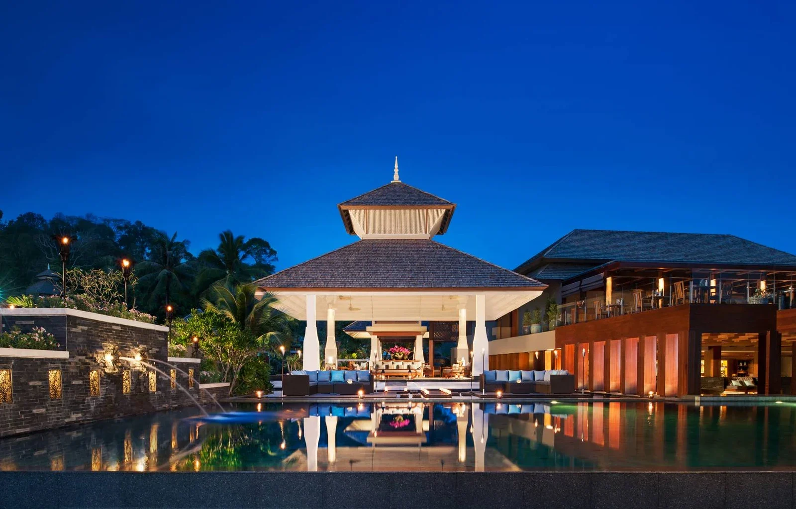 Общий бассейн Anantara Layan Phuket Resort отражает освещённый павильон и соседнее здание на фоне вечернего неба.