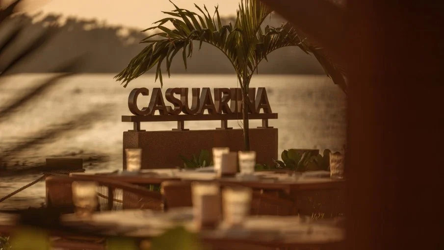 Вывеска Casuarina Beach Club за столиками и пальмами на вечерней террасе