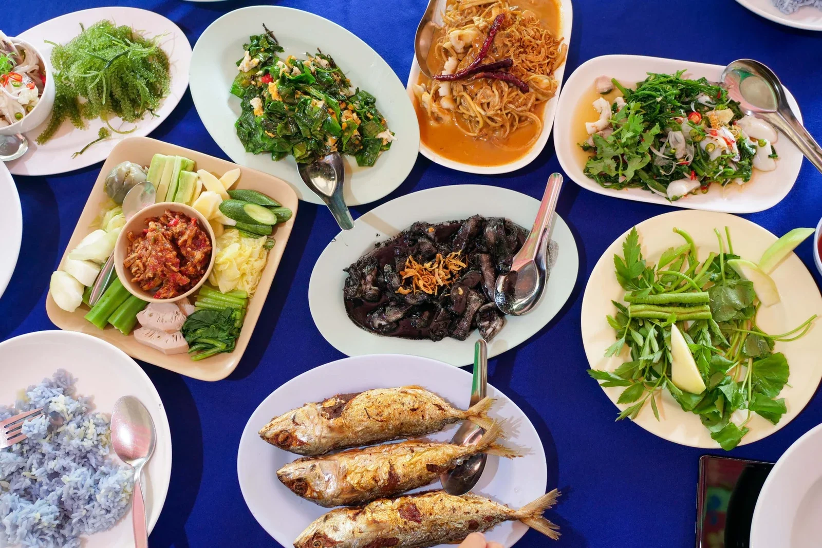 Стол на синей скатерти с блюдами южнотайской кухни: жареная макрель, морской виноград, острые салаты и кальмары