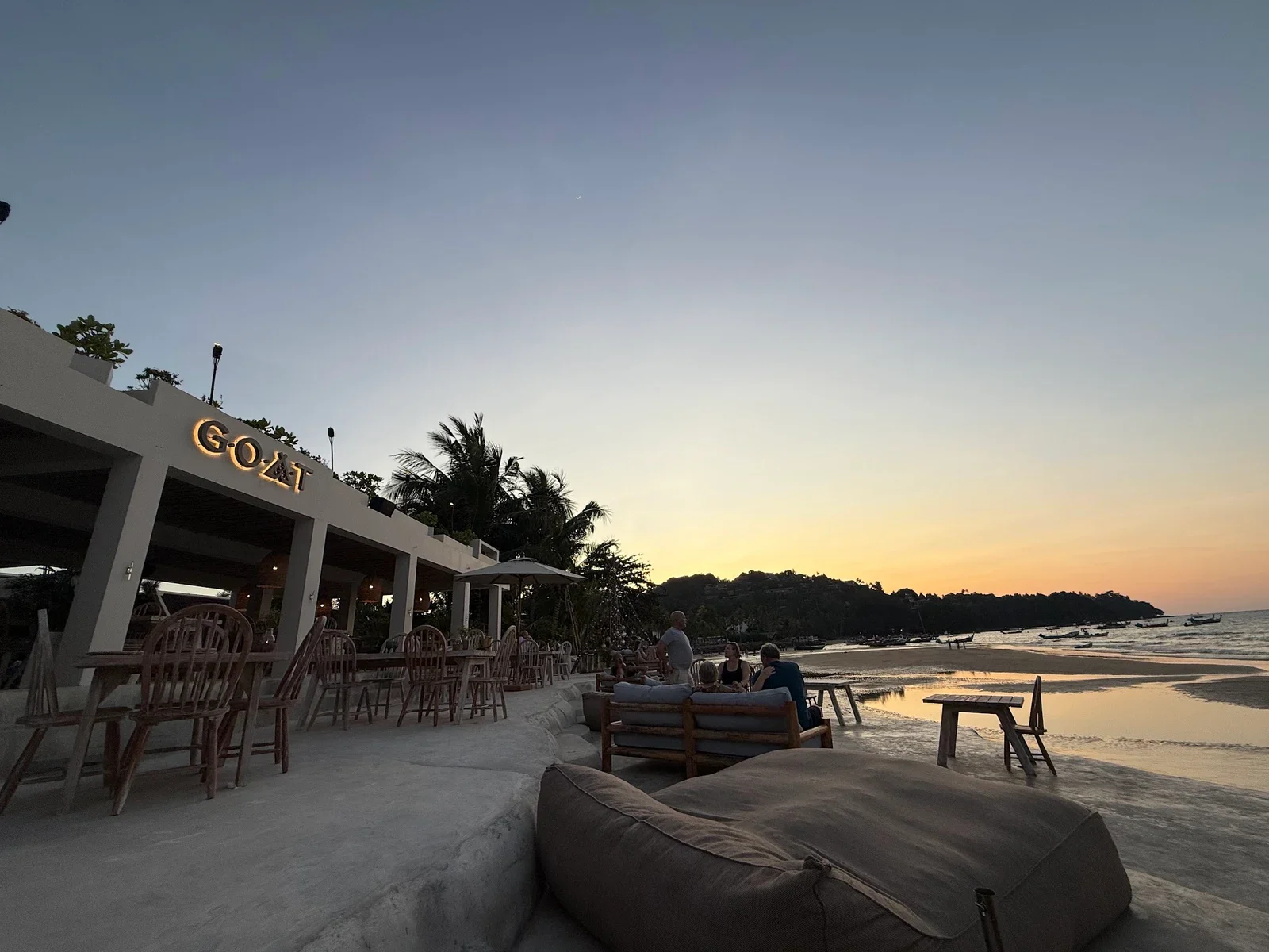 Терраса GOAT Beach Club на закате: светящаяся вывеска, деревянные столы и диваны у кромки воды
