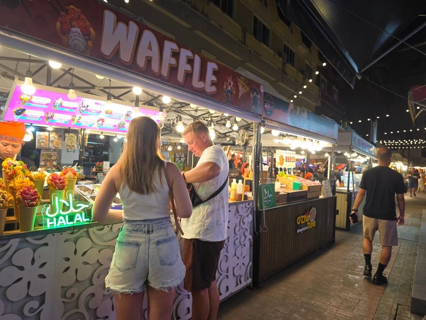 Посетители покупают вафли с фруктами у освещённого прилавка Kata Walking Street вечером