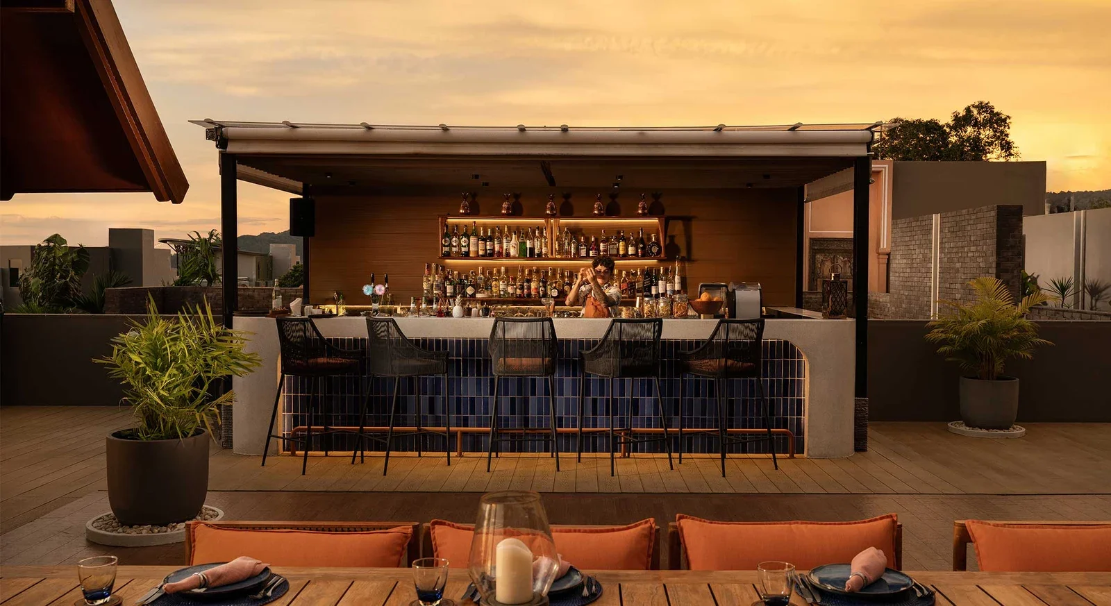 Барная стойка Sizzle Rooftop с высокими стульями и бутылками, бармен готовит напиток в закатный час