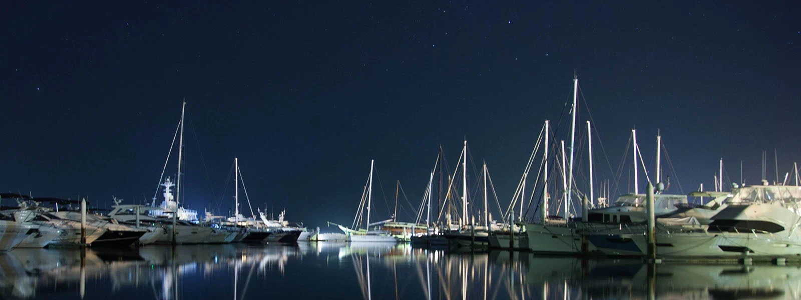 Мачты яхт Ao Po Grand Marina ночью отражаются в тихой воде под звёздным небом.