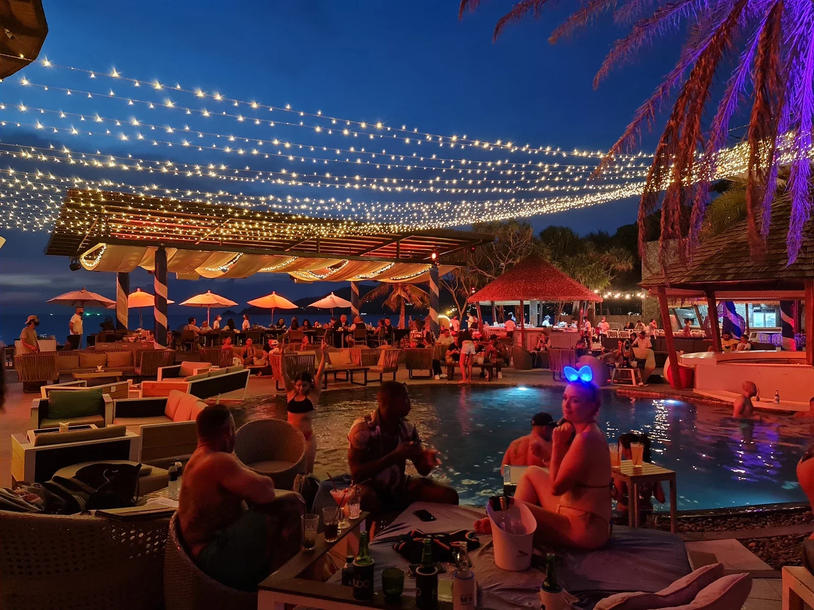Вечерний бассейн KUDO Beach Club: гирлянды над лаунж-зоной, гости на диванах, подсветка воды и пальм