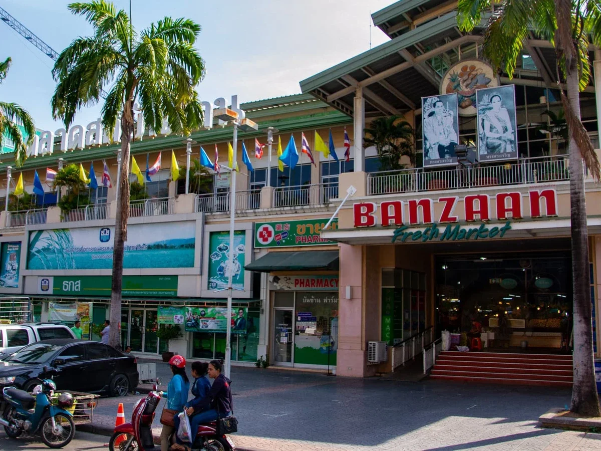 Вход на рынок Banzaan Fresh Market в Патонге: вывеска, флаги, пальмы, припаркованные машины и байки