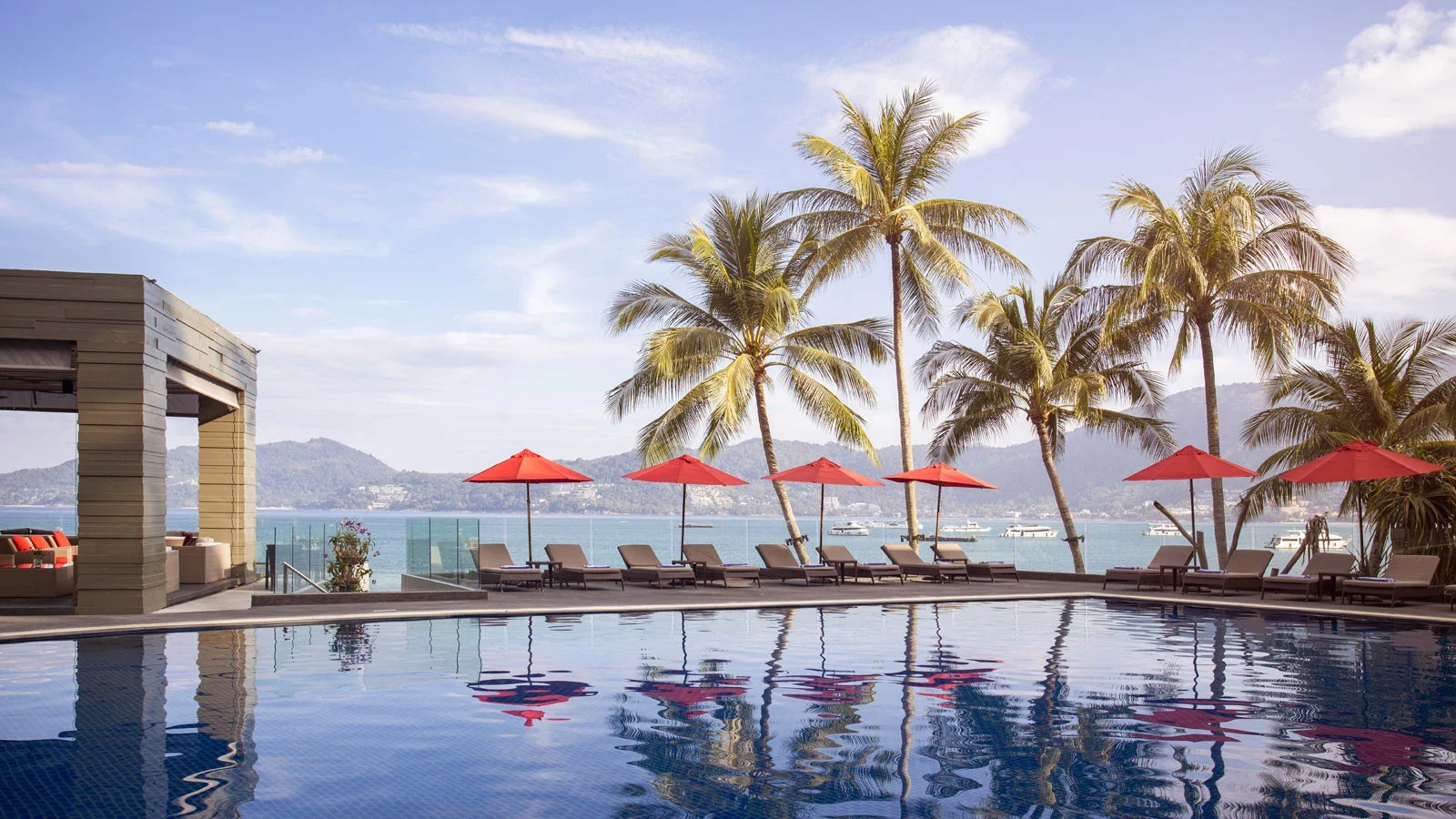 Основной бассейн отеля Amari Phuket с отражением пальм, шезлонги и красные зонты на террасе у залива Патонга