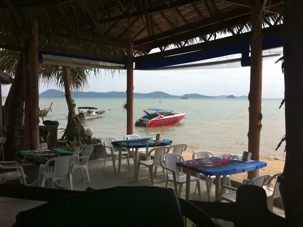 Столики под соломенной крышей пляжного кафе Friendship Beach Resort на песке, за ними залив и катера
