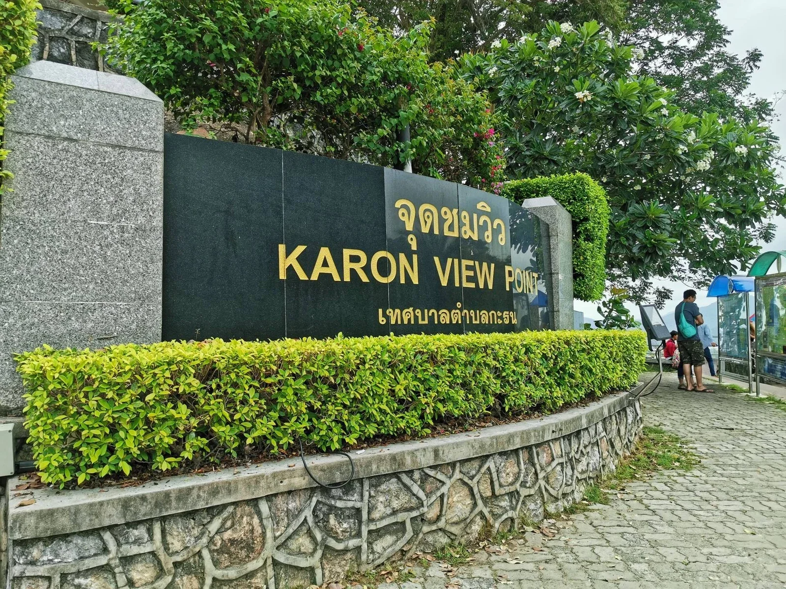 Входная табличка KARON VIEW POINT (จุดชมวิว) на каменной стенке под живой изгородью, у площадки люди