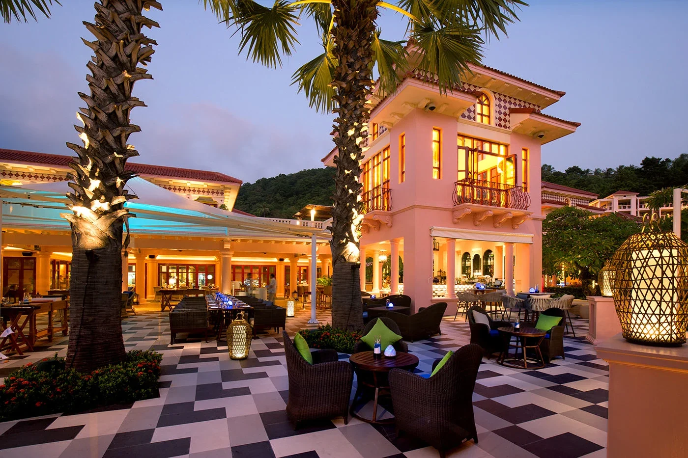Вечерняя терраса Coast Beach Club: подсветка пальм, розовое здание с балконом и столики на шахматной плитке