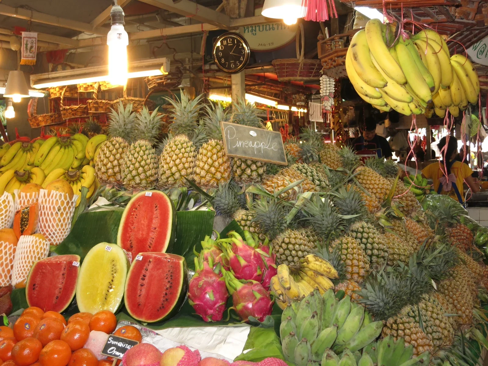 Фруктовый прилавок Banzaan Fresh Market: ананасы, арбузы, питайя, бананы под лампами
