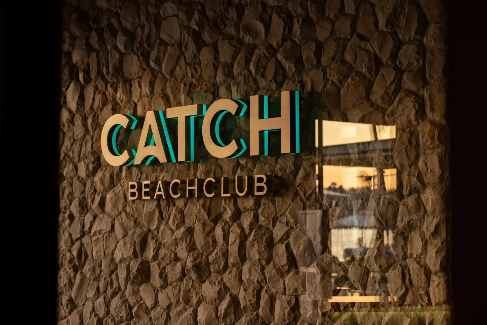 Подсвеченная вывеска CATCH Beach Club на стене из природного камня, вечерний свет