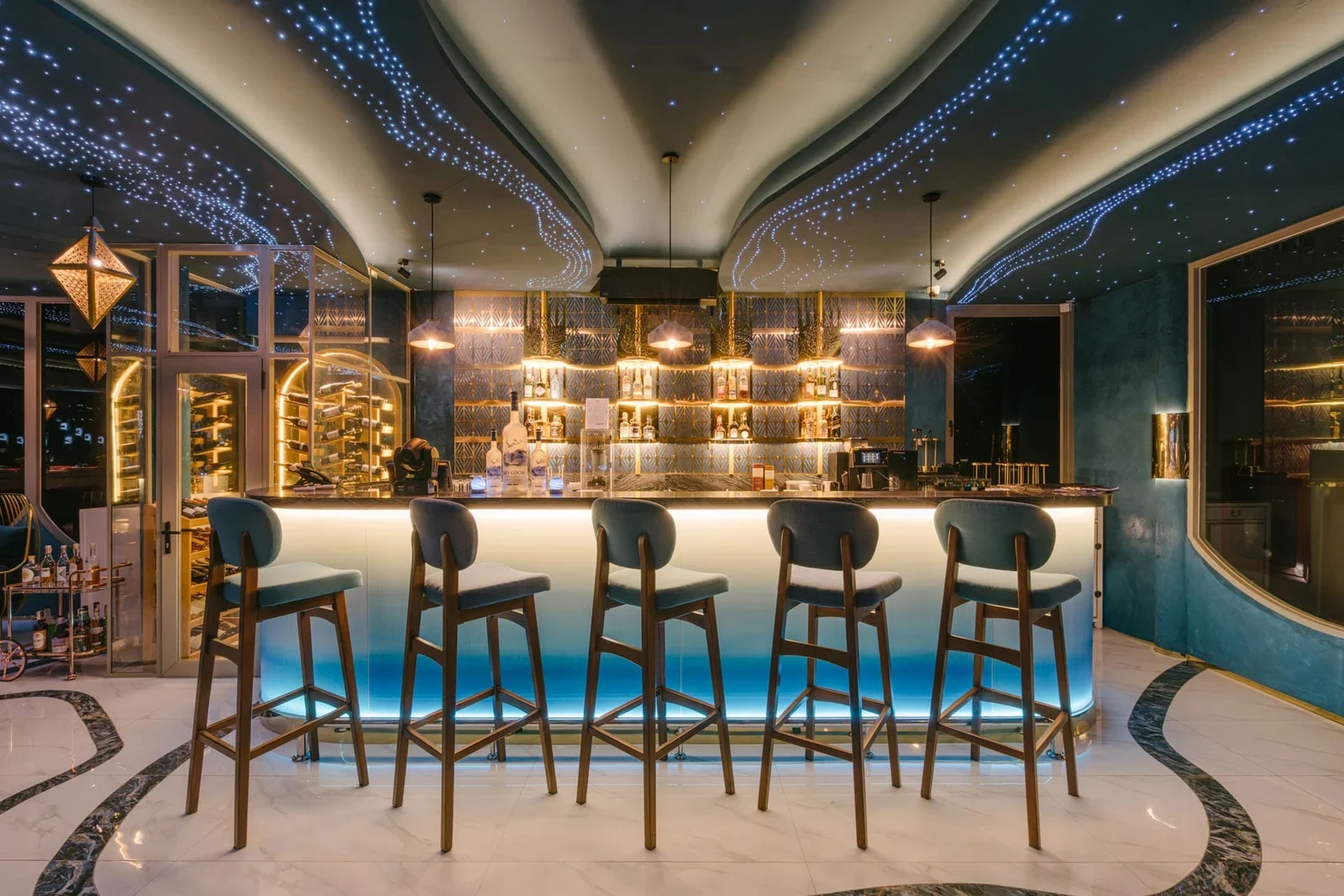 Барная стойка Moon Terrace Lounge с голубой подсветкой, высокие стулья, полки с бутылками и потолок со световыми линиями