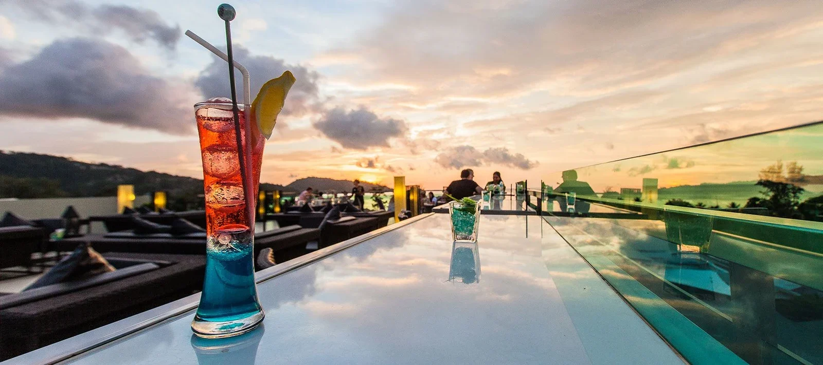 Яркий коктейль на стеклянной стойке крыши Kee Sky Lounge, вдали гости и закатное небо