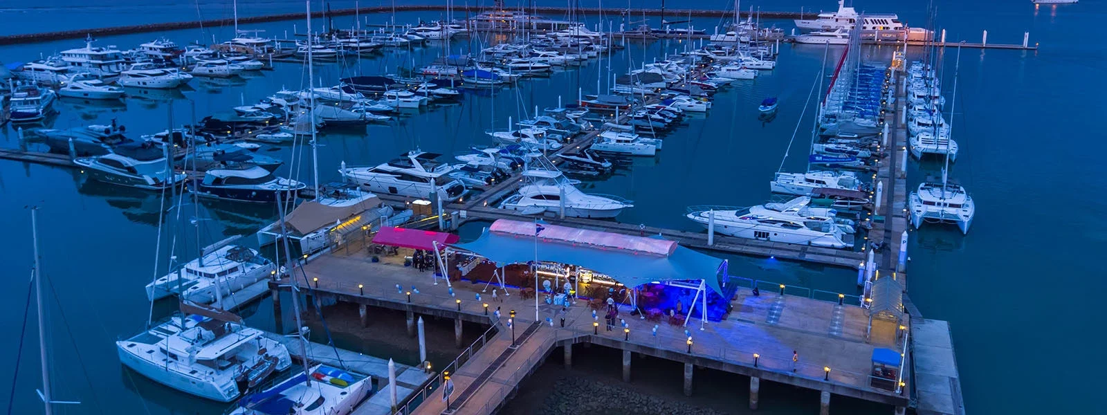 Вечером на пирсе Ao Po Grand Marina освещена зона с тентами среди яхт и катамаранов.