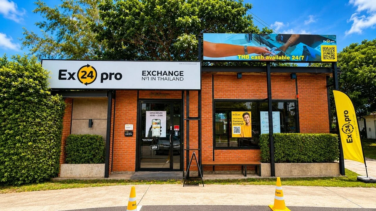Фасад пункта обмена Ex24.pro в Банг Тао: вывеска Exchange целиком, кирпичная стена, жёлтый флаг и оранжевые конусы у входа
