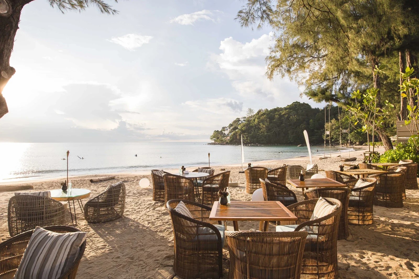 Столики и плетёные кресла на песке пляжа Камала у Pine Beach Bar, вдали лесистый мыс и мягкий закатный свет