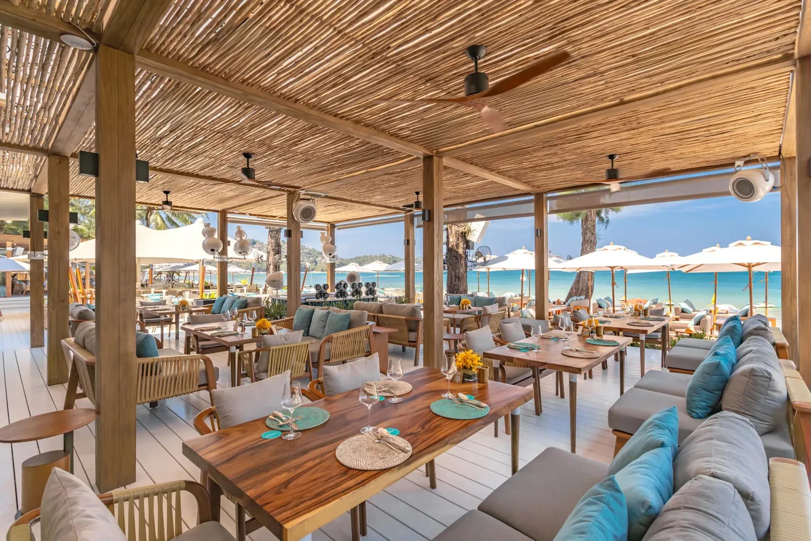 Ресторанный павильон Catch Beach Club с тростниковой крышей, столами и диванами, за ними пляж и вода