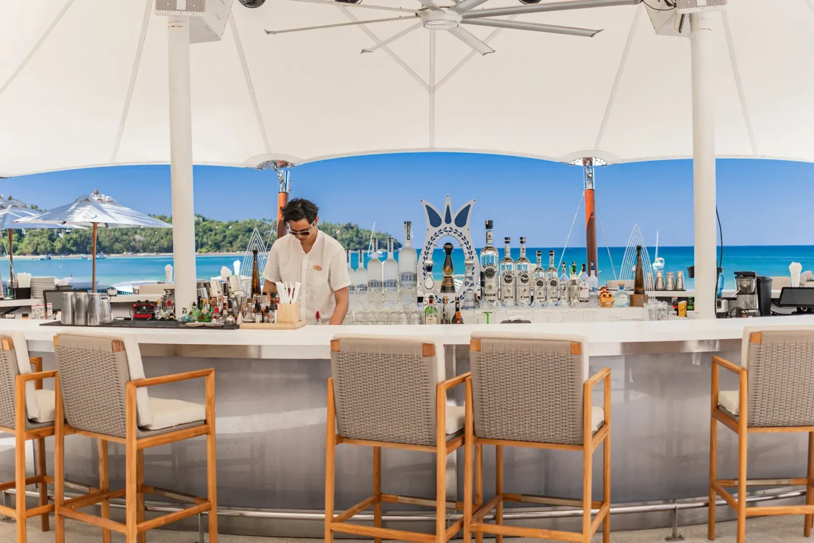 Круглая барная стойка Catch Beach Club под белым тентом, за ней бармен и бирюзовое море