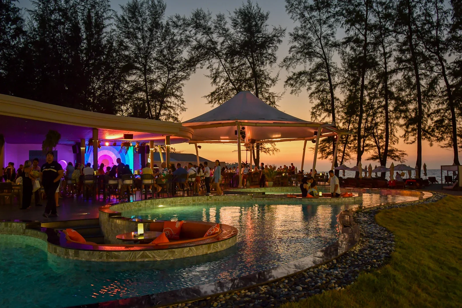 Вечерняя площадка NORA Beach Club с бассейном и подсвеченным баром под деревьями